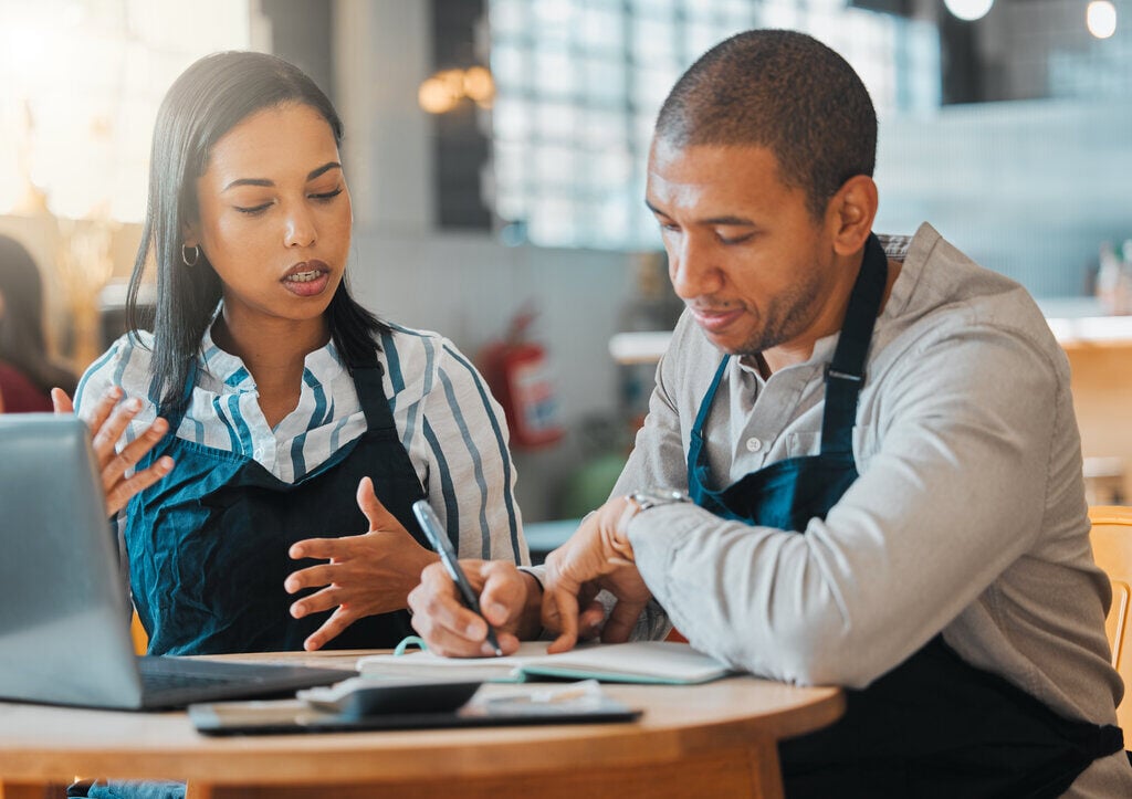 Two owners of a restaurant sit together to work on a laptop and discuss how to save more money for their business.